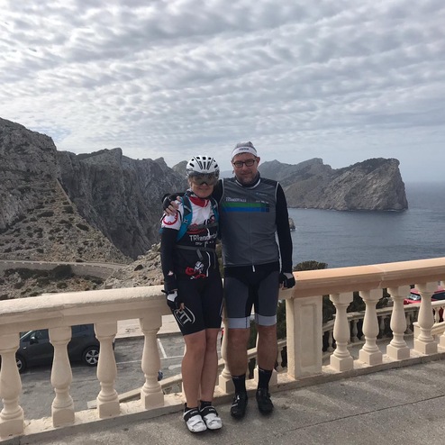 Zwei Radfahrer posieren vor einer Küstenlandschaft mit Bergen und Meer, umgeben von bewölktem Himmel.