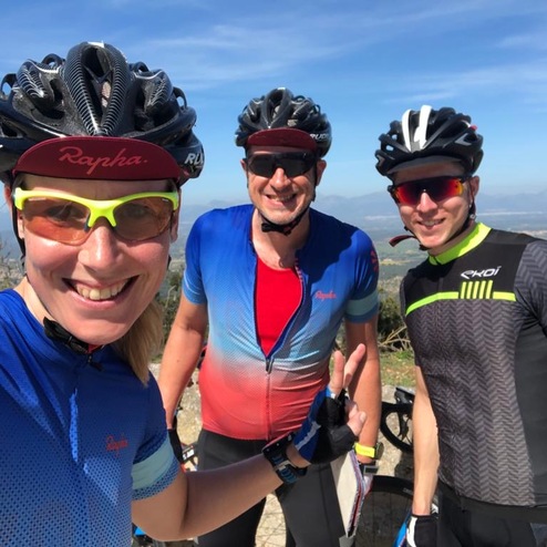 Drei Radfahrer in Sportkleidung stehen auf einem Hügel mit Blick auf die Landschaft, lächeln in die Kamera.