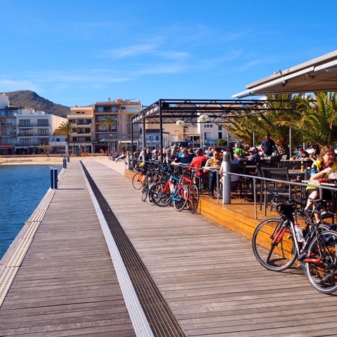 Blick auf eine Holzterrasse am Wasser mit Sitzplätzen, Fahrrädern und Gästen bei schönem Wetter.