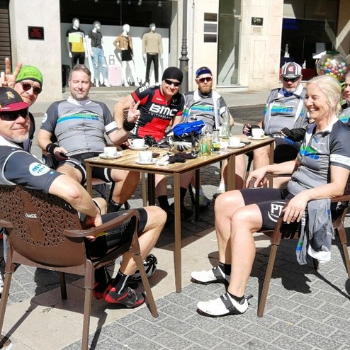 Gruppe von sieben Radfahrern in Sportkleidung sitzt an einem Tisch im Freien, Getränken und Speisen vor ihnen.