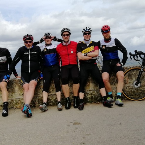 Sechs Radfahrer in Sportkleidung sitzen auf einer Mauer, ein Fahrrad steht daneben, Hintergrund mit Wolken.