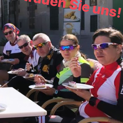 Fünf Radfahrer in Sportkleidung genießen Kuchen und Getränke im Freien vor einer Stadtansicht.