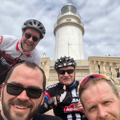 Vier Radfahrer posieren lächelnd vor einem Leuchtturm unter bewölktem Himmel.