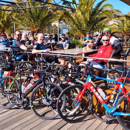 Gruppe von Radfahrern sitzt entspannt im Freien, mit mehreren Fahrrädern vor einem Restaurant unter Palmen.