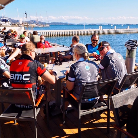 Gruppe von Radfahrern in Sportbekleidung sitzt an einem Tisch am Wasser und genießt die Aussicht.