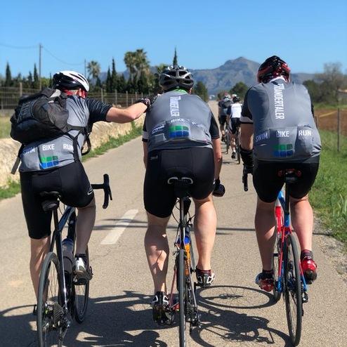Drei Radfahrer in Sportkleidung fahren auf einer Straße in einer landschaftlichen Umgebung.