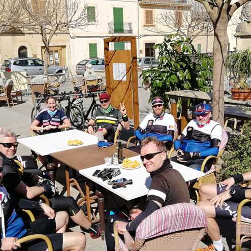 Gruppe von Radfahrern sitzt entspannt in einem Café unter Bäumen, Tisch mit Snacks und Getränken in der Mitte.