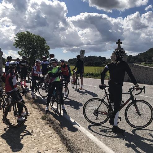 Gruppe von Radfahrern hält an einer Straße mit einer Steinkreuzung und grüner Landschaft im Hintergrund.