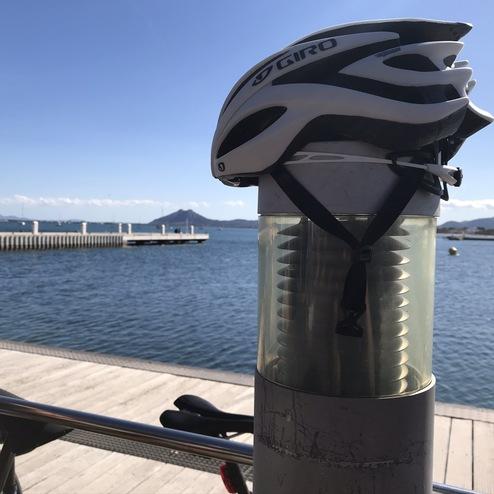 Helm auf einem Poller, dahinter ein Hafen mit Holzsteg und Wasser, Berge am Horizont unter blauem Himmel.