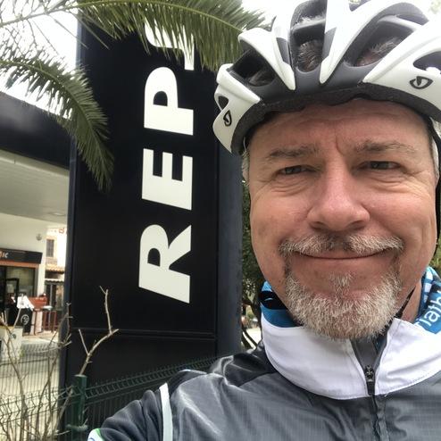 Selbstporträt eines Radfahrers mit Helm vor einem großen „REPSOL“-Schild und Palmen im Hintergrund.