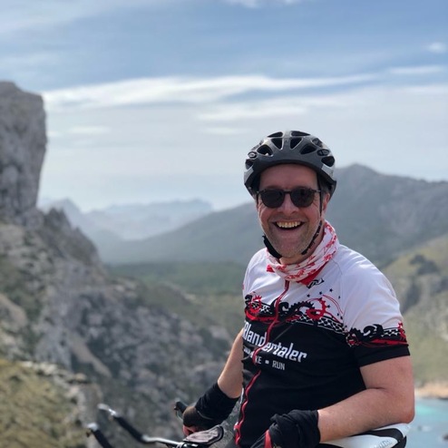 Lächelnder Radfahrer in Fahrradbekleidung sitzt auf seinem Rennrad mit Berglandschaft im Hintergrund.