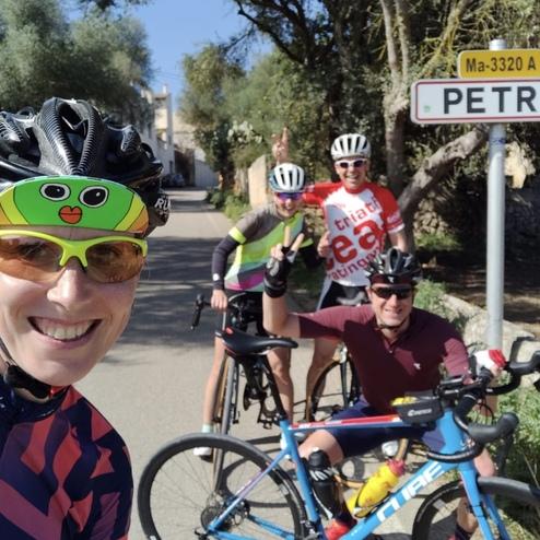 Gruppe von vier Radfahrern posiert lächelnd vor einem "Petra"-Ortsschild auf einer Straße.