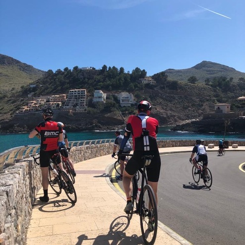 Gruppe von Radfahrern auf einer Uferpromenade mit Aussicht auf das Meer und umliegende Hügel.