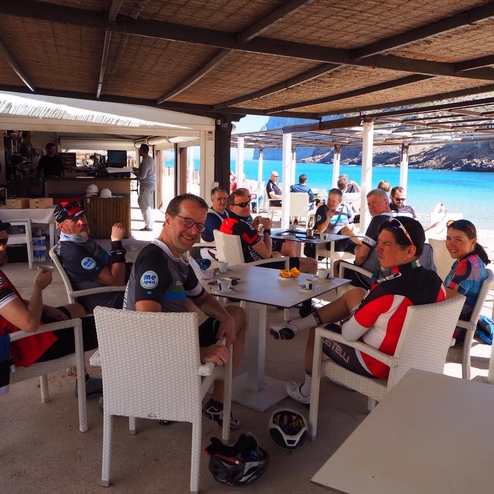 Gruppe von Radfahrern sitzt entspannt unter einem Sonnenschutz in einem Strandcafé mit Meerblick.