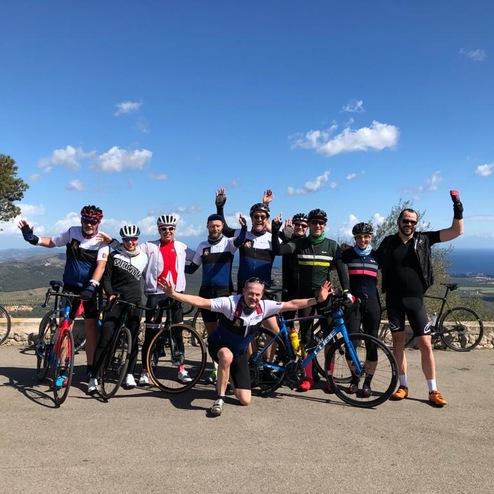 Gruppe von zehn Radfahrern posiert fröhlich vor einer Landschaft mit Meerblick und mehreren Fahrrädern im Hintergrund.