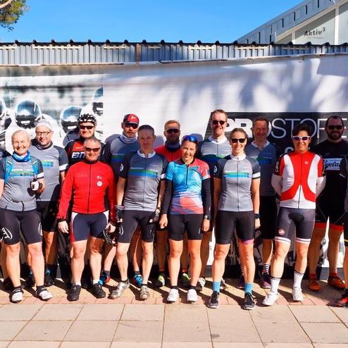 Gruppe von Radfahrern in Sportbekleidung posiert vor einem Veranstaltungsbanner an einem sonnigen Tag.