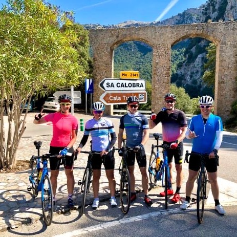 Gruppe von fünf Radfahrern posiert mit Fahrrädern vor einem historischen Bogen und Verkehrszeichen in der Landschaft.