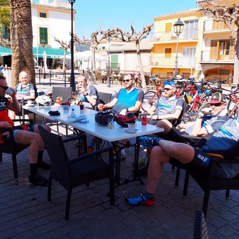 Gruppe von Radfahrern sitzt an einem Tisch in einem Café, umgeben von Fahrrädern und Palmen.