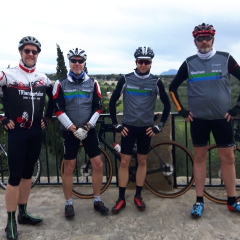 Vier Radfahrer in Sportbekleidung posieren mit ihren Fahrrädern vor einer grünen Landschaft unter bewölktem Himmel.
