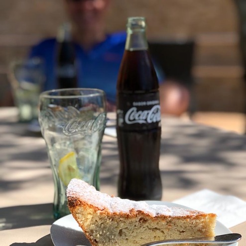 Stück Kuchen mit Puderzucker auf einem weißen Teller, ein Glas und eine Flasche Coca-Cola im Hintergrund.
