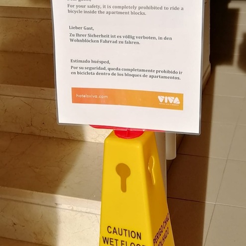 Gelbes Warnschild mit Texten in mehreren Sprachen, das vor rutschigem Boden und dem Verbot, mit dem Fahrrad zu fahren, warnt.