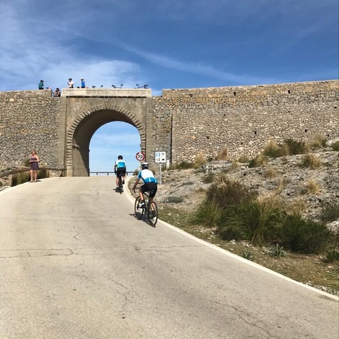 Radfahrer fahren eine steile Straße unter einem großen, steinernen Bogen, umgeben von Gestrüpp und Sand.