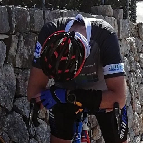 Radfahrer in Sportbekleidung beugt sich müde über sein Fahrrad vor einer Steinmauer.