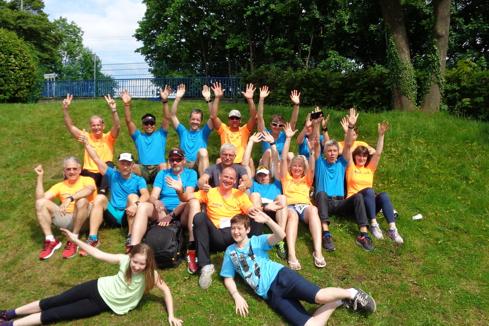 Gruppensnapshot von 20 Personen in bunten T-Shirts, sitzend auf einer Wiese, alle lächeln und heben die Hände.