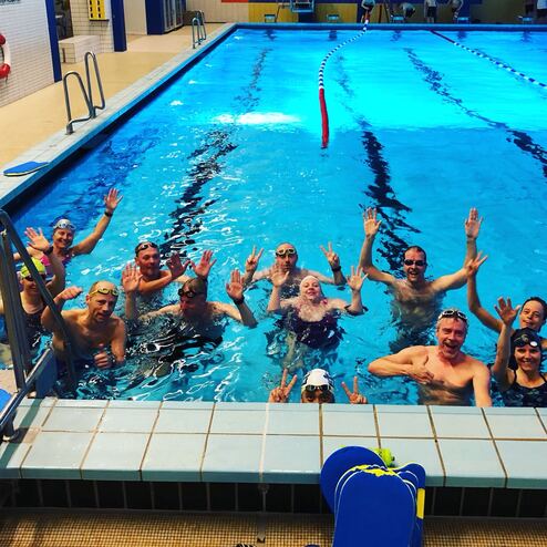 Gruppe von Schwimmern im Wasser, die freundlich in die Kamera winken und posieren, hinter ihnen ein Schwimmbad.