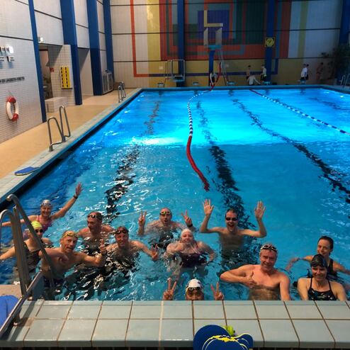 Gruppenszene mit Schwimmern im Wasser, die lächeln und winken, in einem hellen Sportbad mit blauer Wasseroberfläche.