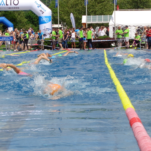 Schwimmer in bunten Badeanzügen absolvieren ein Rennen in einem Pool, begleitet von Zuschauern am Beckenrand.