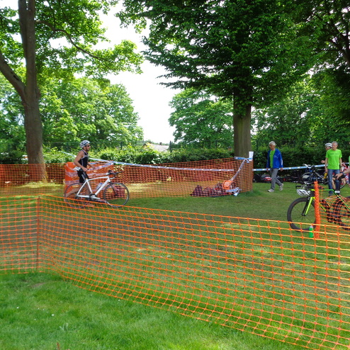 Teilnehmer eines Triathlons bewegen sich an einer gesperrten Fläche mit orangem Absperrnetz und Fahrrädern im Hintergrund.