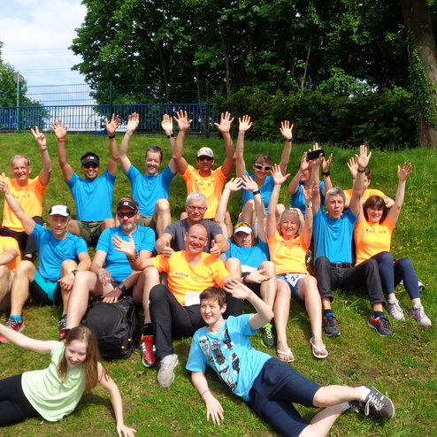 Gruppensnapshot von 20 Personen in bunten T-Shirts, sitzend auf einer Wiese, alle lächeln und heben die Hände.