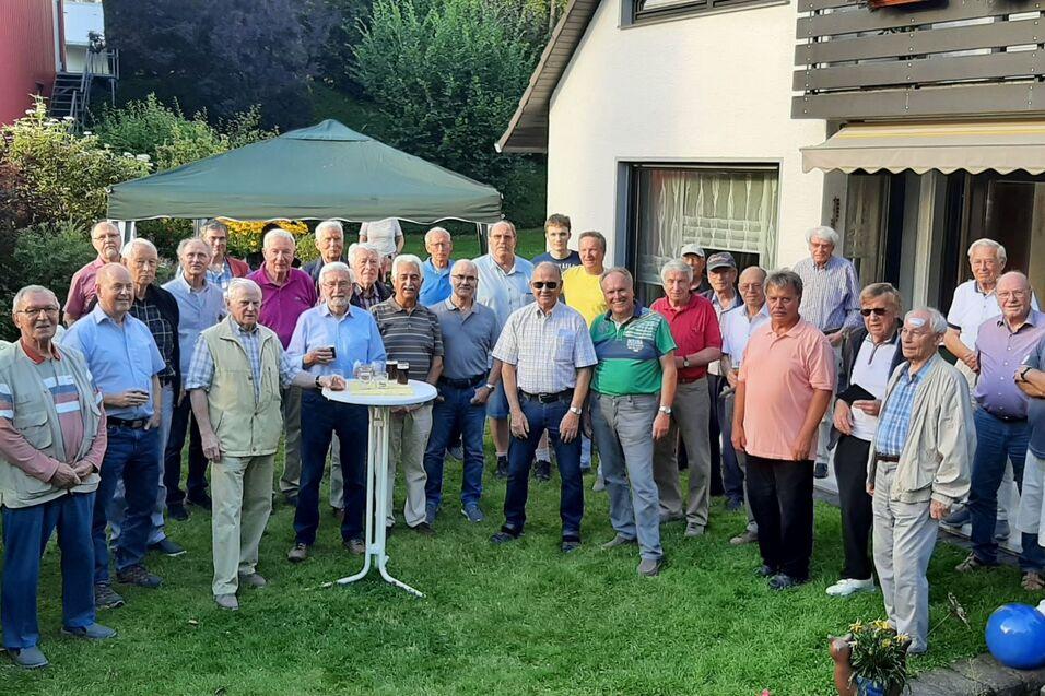 Gruppe von Männern in einem Garten, einige halten Gläser, steht vor einem Haus mit grüner Rasenfläche und Blumen.