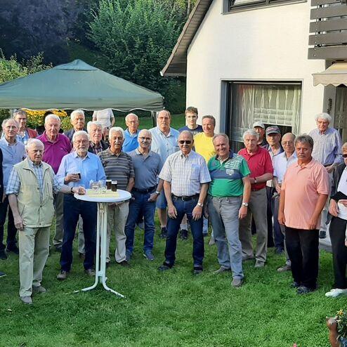 Gruppe von Männern in einem Garten, einige halten Gläser, steht vor einem Haus mit grüner Rasenfläche und Blumen.