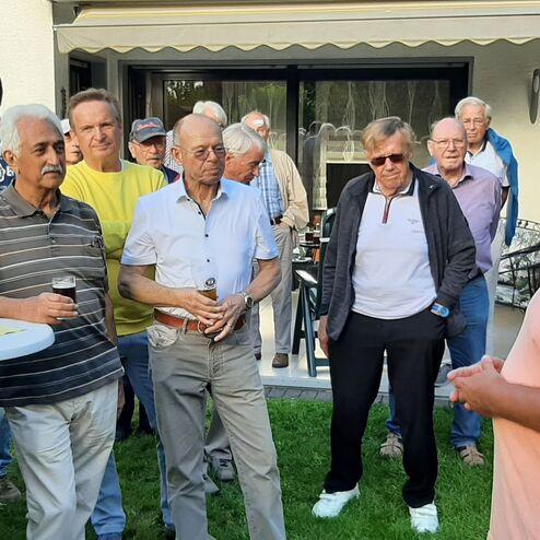 Eine Gruppe älterer Männer hört einem Redner im Freien aufmerksam zu, einige halten Getränke in der Hand.