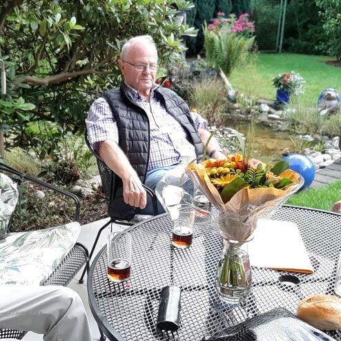 Drei ältere Männer sitzen an einem Tisch im Garten, auf dem sich Getränke und ein Blumenstrauß befinden.