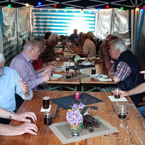 Gruppenszene mit älteren Männern, die an einem Tisch essen und sich unterhalten, unter einem Zeltdach mit Lichtern.