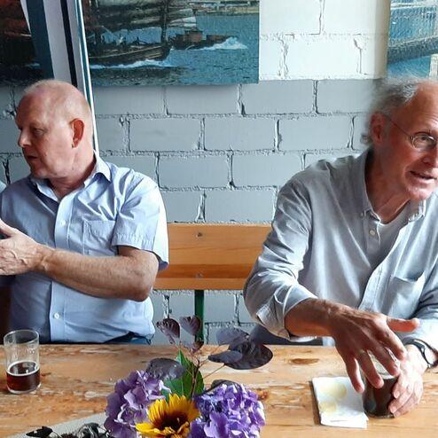 Vier Männer sitzen an einem Tisch, unterhalten sich, mit Biergläsern und einer Blumendekoration vor ihnen.