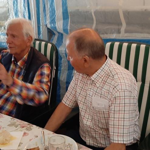 Zwei ältere Männer unterhalten sich an einem Tisch, im Hintergrund eine transparente, gestreifte Abtrennung.