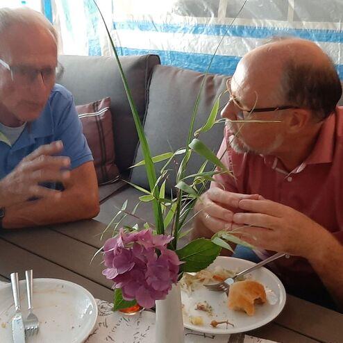 Zwei Männer sitzen an einem Tisch, essen und unterhalten sich, während eine Blume in der Mitte steht.