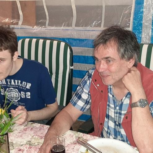 Zwei Personen sitzen an einem Tisch unter einem grünen und blauen Zelt, genießen Essen und trinken aus Gläsern.