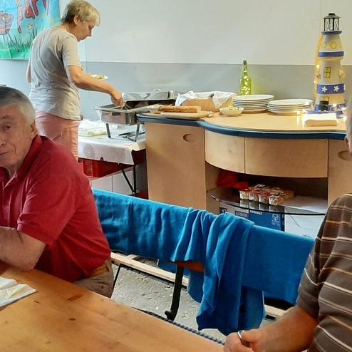 Festliches Beisammensein von älteren Männern an einem Tisch, während eine Frau im Hintergrund Essen serviert.