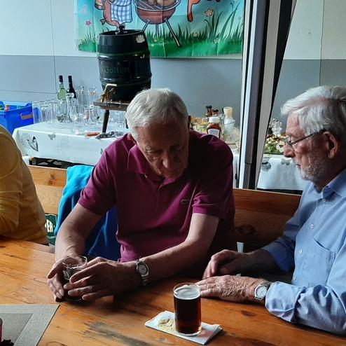 Drei ältere Männer sitzen an einem Holztisch, trinken Bier und unterhalten sich, im Hintergrund Tisch mit Speisen.