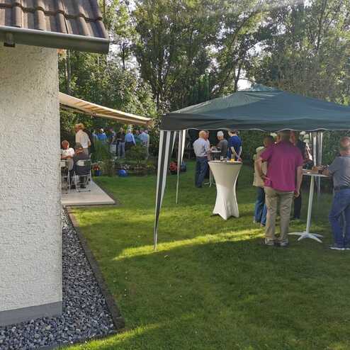 Gruppierung von Personen unter einem Zelt in einem Garten, umgeben von Grünflächen und einem Nebengebäude.