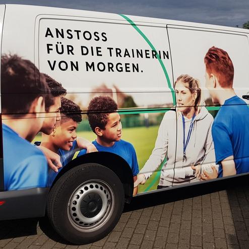 Seitenansicht eines Vans mit einem Motiv für Trainerinnen, darunter Kinder in blauen Trikots auf einem Fußballfeld.