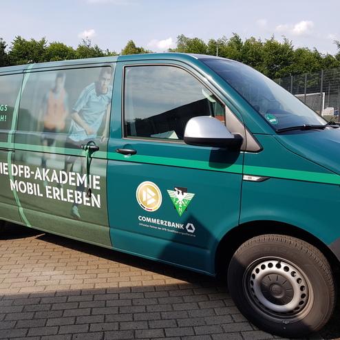 Grüner Transporter mit Werbung für die DFB-Akademie, umgeben von Fußballplätzen und Bäumen im Hintergrund.