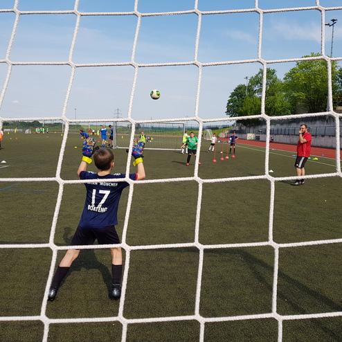 Jungtorwart in Handschuhen steht bereit im Tor, während ein Ball auf ihn zugeschossen wird. Trainingssituation auf Rasenplatz.