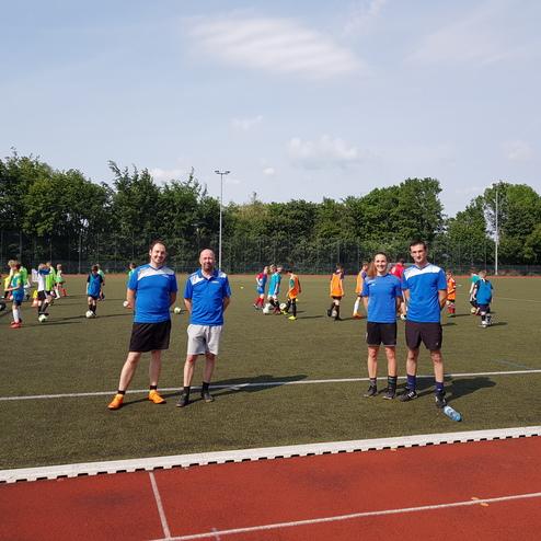 Vier Trainer in blauen Trikots stehen auf einem Kunstrasenplatz, während im Hintergrund Kinder Fußball trainieren.