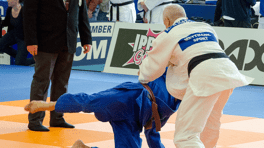 Zwei Judoka im Wettkampf auf einer orangefarbenen Matte, ein Schiedsrichter beobachtet das Geschehen im Hintergrund.